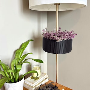 May include: A gold floor lamp with a white lampshade and a black planter hanging from the pole. The planter has a purple and green plant inside. The lamp is next to a wooden bedside table with a white ceramic vase with a green plant, a stack of books, a candle, a pair of glasses, a notebook, a pen, and a hair clip.