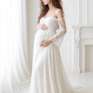 May include: A white, off-the-shoulder maternity gown with a flowing skirt and lace sleeves. The dress is floor-length and features a sweetheart neckline. The model is standing near a window with white curtains.