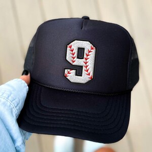 Könnte beinhalten: Marineblaue Trucker-Cap mit einer weiß-roten, gestickten Baseball-Nummer 9. Die Kappe hat einen Mesh-Rücken und einen gebogenen Schirm. Die Nummer 9 ist weiß umrandet und mit roten Nähten versehen.