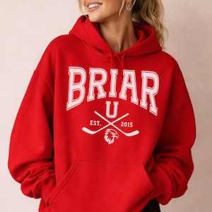 Op de afbeelding: Een rode hoodie met het woord "BRIAR" in witte hoofdletters. Daaronder staat de letter "U", gekruiste hockeysticks, een wapen en de tekst "EST. 2015". De hoodie heeft een capuchon en een voorzak.