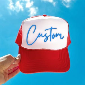 Może przedstawiać: Czerwono-biała czapka typu trucker z napisem "Custom" w niebieskim skrypcie. Czapka jest trzymana na tle jasnoniebieskiego nieba z białymi chmurami. Czapka ma czerwony daszek i siatkową tylną część.