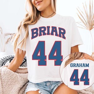 Puede incluir: Camiseta blanca con el nombre "BRIAR" y el número "44" en azul con ribete rojo. Una imagen más pequeña muestra el nombre "GRAHAM" y el número "44" en la espalda. La camiseta es de manga corta.