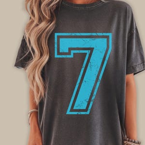 May include: Dark gray t-shirt featuring a large, distressed number 7 in light blue with a black outline. The shirt has a vintage, worn look. The t-shirt is a casual, unisex garment.