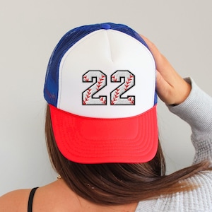 Può includere: Cappellino da baseball in stile trucker con un pannello frontale bianco con il numero "22" in un design a punto baseball. Il cappellino ha una visiera rossa, un retro in rete blu e un bottone blu.