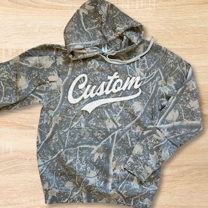 Peut inclure: Un sweat-shirt à capuche à manches longues avec un motif camouflage de branches et de feuilles marron, gris et vert. Le mot Custom est imprimé en écriture blanche au centre de la poitrine. Le sweat inclut une poche avant et des cordons.