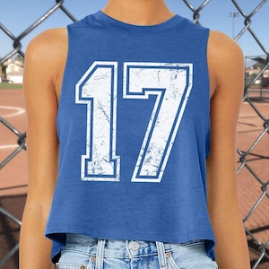 May include: A blue crop top with the number 17 in white, featuring a distressed, vintage-style design. The top is sleeveless and has a round neckline. The background includes a chain-link fence and a baseball field.