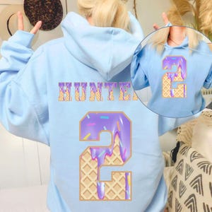Op de afbeelding: Lichtblauwe hoodie met de naam "HUNTER" in een wafelvormig lettertype. Een groot nummer "2" staat eronder, ontworpen als een paarse donut met hagelslag en wafeltextuurdetails. De hoodie heeft een capuchon met trekkoord.