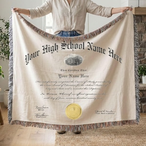 Op de afbeelding: Een crèmekleurige geweven sprei met een diploma-ontwerp. De deken heeft de tekst "Your High School Name Here" en "Your Name Here" in een vintage lettertype. De randen hebben een franjebeslag.