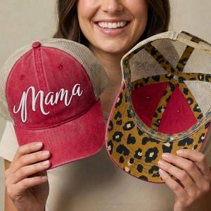May include: Two red trucker hats. One hat has the word "Mama" embroidered in white script. The other hat has a leopard print design on the underside of the brim. Both hats have a mesh back and a curved brim.