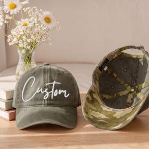 May include: Two baseball caps, one olive green with the word Custom embroidered in white script on the front, and one showing the interior with a green and brown camouflage pattern on the underside of the brim and inner lining panels.