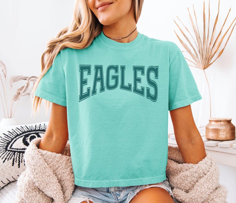 May include: A short-sleeved, crew-neck t-shirt in a bright turquoise color. The front of the shirt features the word "EAGLES" printed in a dark teal, arched, collegiate-style font with a double-line outline effect across the chest area.