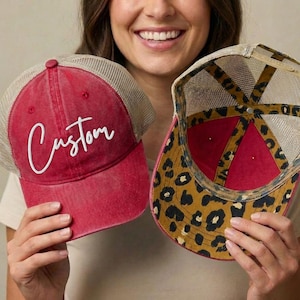 May include: Two red trucker hats with mesh backs are held up. One hat has the word "Custom" in white script. The other hat has a leopard print pattern on the underside of the brim. Both hats have a red crown.