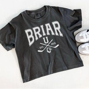 May include: Dark gray t-shirt with the word "BRIAR" in white block letters, with "U" below and crossed hockey sticks. Below the sticks is "EST. 2015" and an eagle. White sneakers with black stripes are next to the shirt.