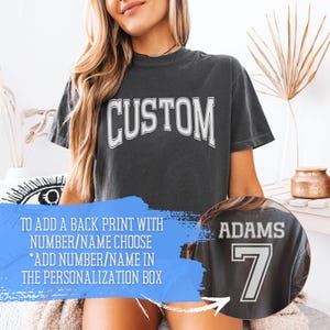 May include: A charcoal gray short-sleeve t-shirt featuring the word CUSTOM in white varsity-style lettering on the front. A circular inset shows the back of the shirt with the name ADAMS and the number 7 in white varsity-style lettering.
