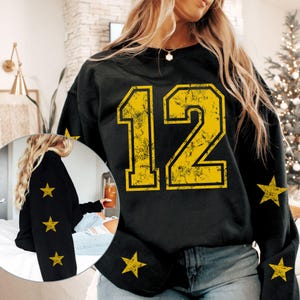 May include: Black sweatshirt with the number 12 in distressed yellow font. The sleeves feature yellow star graphics. The sweatshirt is a casual style, suitable for everyday wear. The design has a vintage or retro aesthetic.