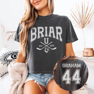 May include: A charcoal gray short-sleeve t-shirt featuring white collegiate-style text that reads BRIAR U, EST. 2015, with crossed hockey sticks and an eagle head icon. The back of the shirt displays the name GRAHAM and the number 44 in white.
