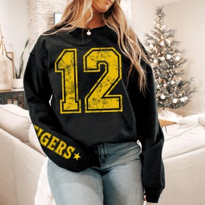 May include: Black sweatshirt with the number 12 in distressed yellow font on the front. The sleeve features the word "TIGERS" in yellow with a star. The sweatshirt is worn by a person.