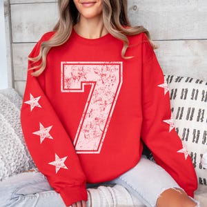 May include: A red sweatshirt featuring a large, distressed white number 7 outlined in red. White stars are printed on the sleeves. The sweatshirt is a crew neck style.