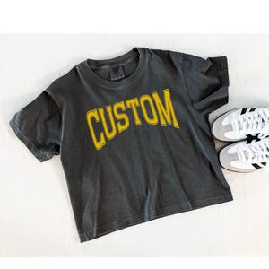 May include: A charcoal grey short-sleeve t-shirt featuring the word CUSTOM printed in a bold, arched yellow font across the chest. The shirt has a crew neckline and a slightly faded, vintage-inspired wash. A pair of white and grey sneakers is visible.