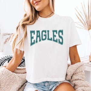 May include: A white short-sleeved t-shirt featuring the word EAGLES printed in a dark teal, arched collegiate-style font with a white outline. The shirt has a crew neckline and a relaxed fit, constructed from a solid white fabric.