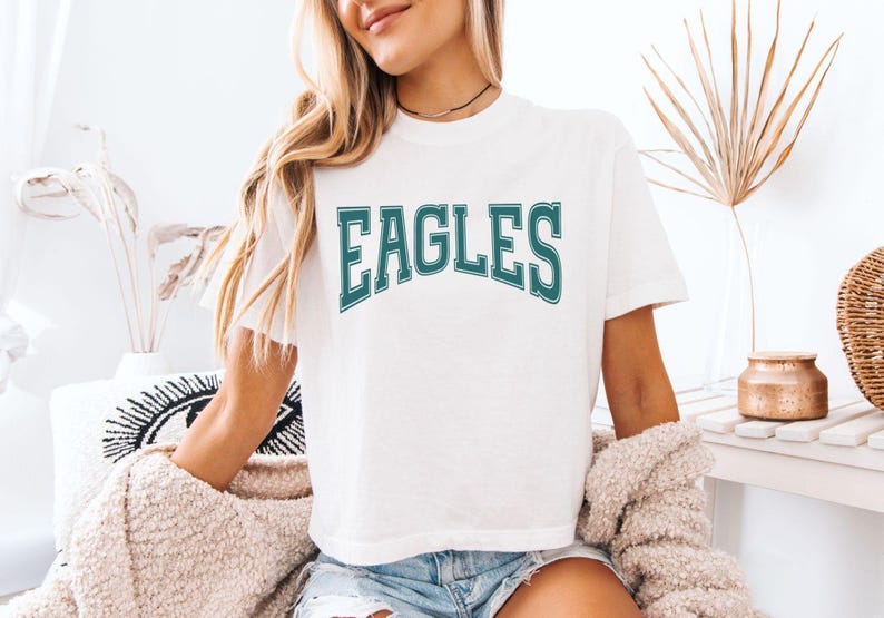 May include: A white short-sleeved t-shirt featuring the word EAGLES printed in a dark teal, arched collegiate-style font with a white outline. The shirt has a crew neckline and a relaxed fit, constructed from a solid white fabric.