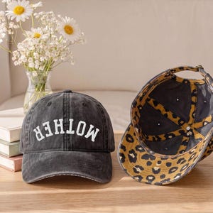 May include: A charcoal grey baseball cap with the word MOTHER embroidered in white on the front. The cap features a leopard print pattern on the underside of the brim and interior panels, with an adjustable metal buckle strap at the back.
