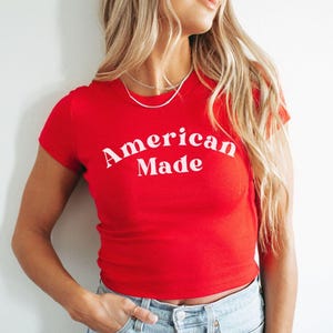 以下が含まれることがあります： 胸元に太字の曲線的なセリフ体でAmerican Madeと白くプリントされた、鮮やかな赤の半袖Tシャツ。この衣類はクラシックなクルーネックと体にフィットするシルエットが特徴で、ライトブルーのデニムジーンズと組み合わされています。