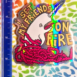 May include: Holographic sticker featuring a cartoon narwhal with a unicorn horn and the text "I SET MY FRIENDS ON FIRE." The narwhal is white with a pink tongue and a pink, rope-like design. The text is in yellow and orange.