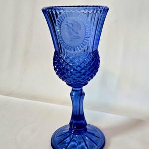 May include: A cobalt blue glass goblet with a diamond pattern and a raised medallion featuring a profile portrait. The goblet has a tall, slender stem and a wide, flared base.