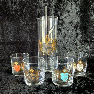 May include: A set of six clear glass cocktail glasses with gold trim and a matching pitcher. Each glass features a different colored crest with a gold lion and a gold crown.