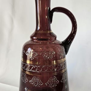May include: A purple glass pitcher with a gold and white floral design. The pitcher has a wide base and a narrow neck with a curved handle.