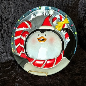 May include: A round glass serving plate with a penguin wearing a red and white striped scarf and a Santa hat. The penguin is holding a candy cane and a yellow bow. The plate has a blue and white background with a snowflake design.
