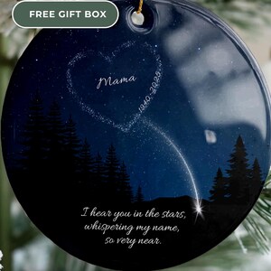 Può includere: Un ornamento rotondo in ceramica blu scuro con un design celeste. Presenta una costellazione a forma di cuore con la parola "Mama" e gli anni "1940-2025". L'ornamento include il testo "I hear you in the stars, whispering my name, so very near."