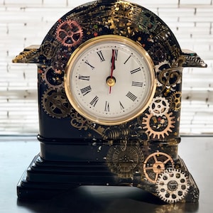 Op de afbeelding: Een zwarte klok met gouden accenten en een wit gezicht. De klok is versierd met tandwielen en andere steampunk-geïnspireerde elementen. De klok staat op een witte bakstenen achtergrond.
