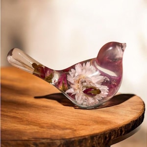May include: A clear, bird-shaped resin figurine containing embedded dried flowers. The sculpture features a large, light-colored central flower surrounded by smaller, deep pink rosebuds, all encased within a smooth, transparent, bird-shaped body.