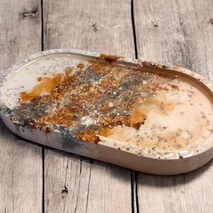 Puede incluir: Una bandeja ovalada con una base blanca y un diseño de resina marrón, dorado y plateado. La resina tiene un efecto marmoleado e incluye pequeñas partículas brillantes.