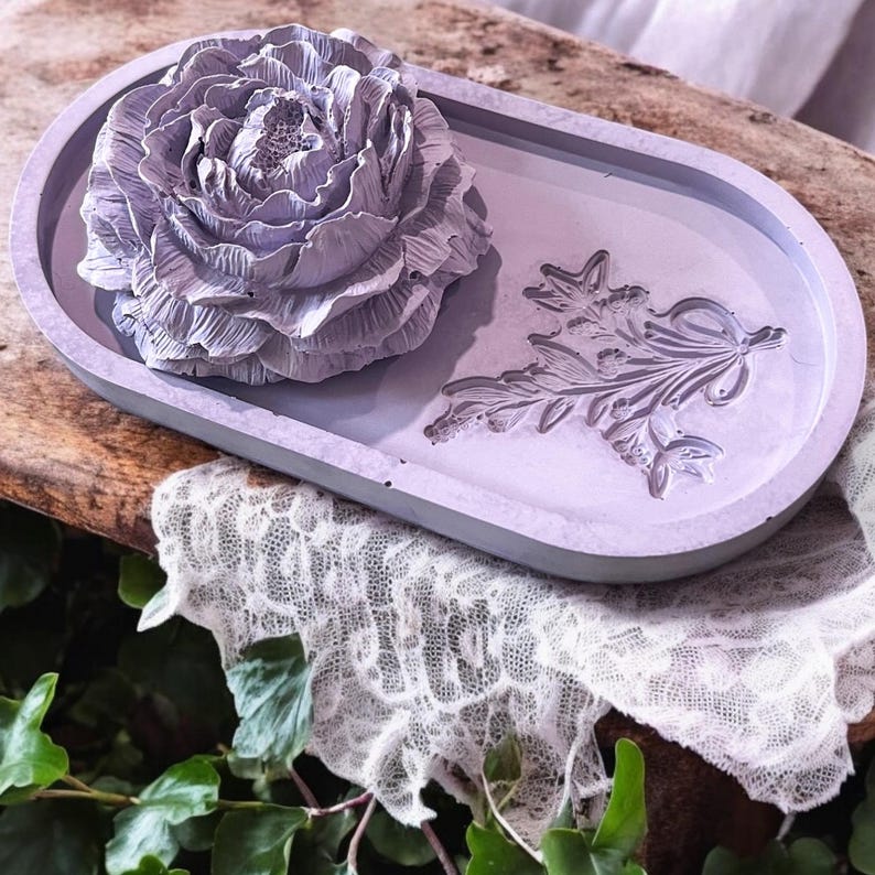 A ceramic embossed oval tray that is lavender and color with a ceramic peony aromatherapy passive diffuser sitting on top of it. They are on a rustic wooden table with a lace cloth, and Ivy is nearby.