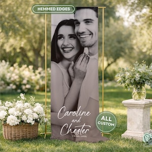 May include: A vertical rectangular banner featuring a black and white photograph of a smiling couple. The text "Caroline and Chester" is printed in white script at the bottom. The banner is suspended from a gold metal stand and has hemmmed edges.