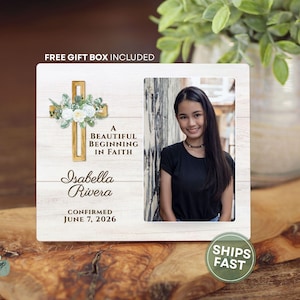 May include: A rectangular white wood-grain plaque featuring a gold cross with white floral accents, the text A BEAUTIFUL BEGINNING IN FAITH, the name Isabella Rivera, and the date CONFIRMED JUNE 7, 2026, alongside a portrait of a young person.