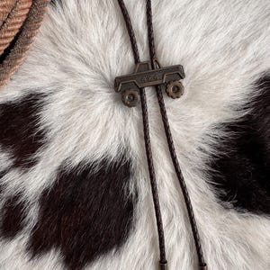 May include: A bolo tie featuring a bronze-toned metal pendant shaped like a 4x4 off-road vehicle. The pendant is attached to a dark brown braided leather cord that ends in two matching bronze-toned metal tips at the bottom of the tie.