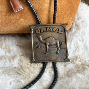 May include: A vintage bolo tie with a rectangular bronze-colored pendant. The pendant features the word "CAMEL" above an image of a camel, palm trees, and a pyramid. The tie has a black braided cord and a brown leather slide.