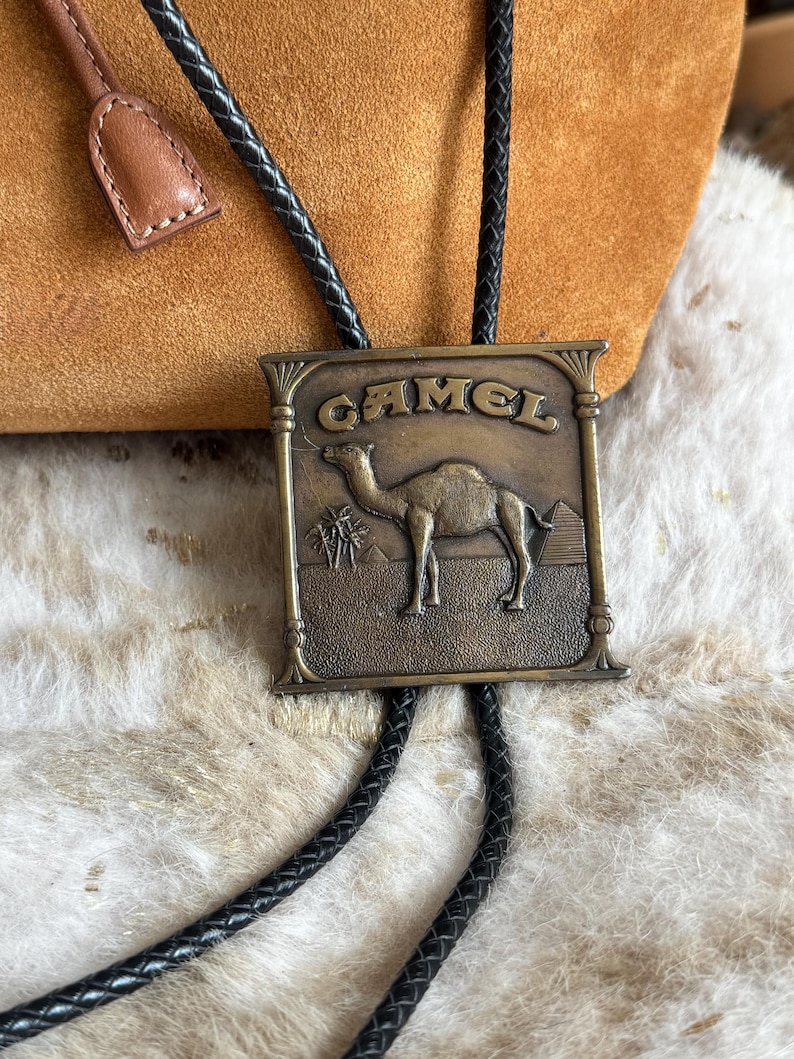 May include: A vintage bolo tie with a rectangular bronze-colored pendant. The pendant features the word "CAMEL" above an image of a camel, palm trees, and a pyramid. The tie has a black braided cord and a brown leather slide.