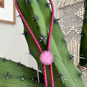 May include: A pink leather bolo tie with a round pink and white agate stone pendant. The bolo tie is wrapped around a cactus.
