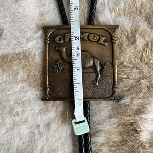 May include: A vintage bolo tie featuring a rectangular brass-toned pendant with the word "CAMEL" and a camel illustration. The pendant is approximately 2 inches (5 cm) in height. The tie has a black braided leather cord.