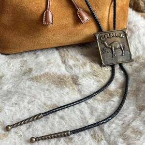 May include: A vintage bolo tie featuring a rectangular bronze-toned metal pendant with the word "CAMEL" and a camel illustration. The tie has a black braided cord with decorative metal tips and a brown suede bag in the background.