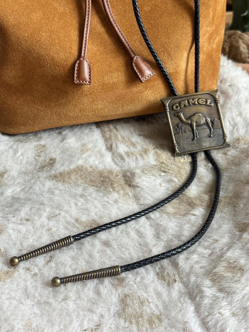 May include: A vintage bolo tie featuring a rectangular bronze-toned metal pendant with the word "CAMEL" and a camel illustration. The tie has a black braided cord with decorative metal tips and a brown suede bag in the background.