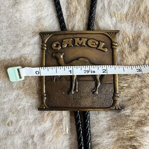 May include: Vintage bolo tie with a rectangular bronze-colored pendant. The pendant features the word "CAMEL" above an image of a camel. The pendant measures approximately 2 inches wide. The tie has a black braided cord.