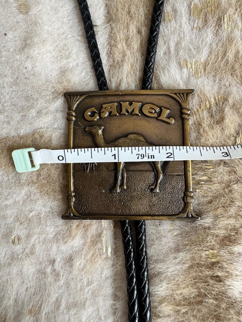 May include: Vintage bolo tie with a rectangular bronze-colored pendant. The pendant features the word "CAMEL" above an image of a camel. The pendant measures approximately 2 inches wide. The tie has a black braided cord.