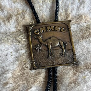 May include: A vintage bolo tie with a rectangular, bronze-colored pendant. The pendant features the word "CAMEL" above a detailed image of a camel in a desert landscape. The tie has a black braided cord.