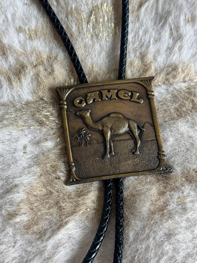 May include: A vintage bolo tie with a rectangular, bronze-colored pendant. The pendant features the word "CAMEL" above a detailed image of a camel in a desert landscape. The tie has a black braided cord.